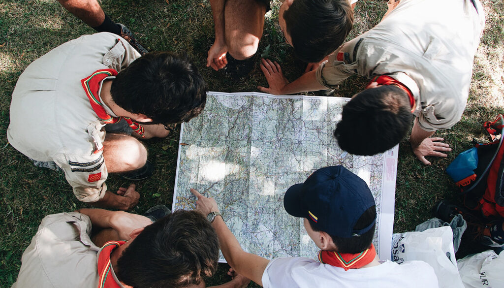 Group of people sitting on grass gathered around a large paper map, pointing and discussing directions.