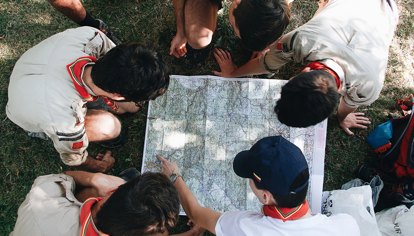 Group of people sitting on grass gathered around a large paper map, pointing and discussing directions.