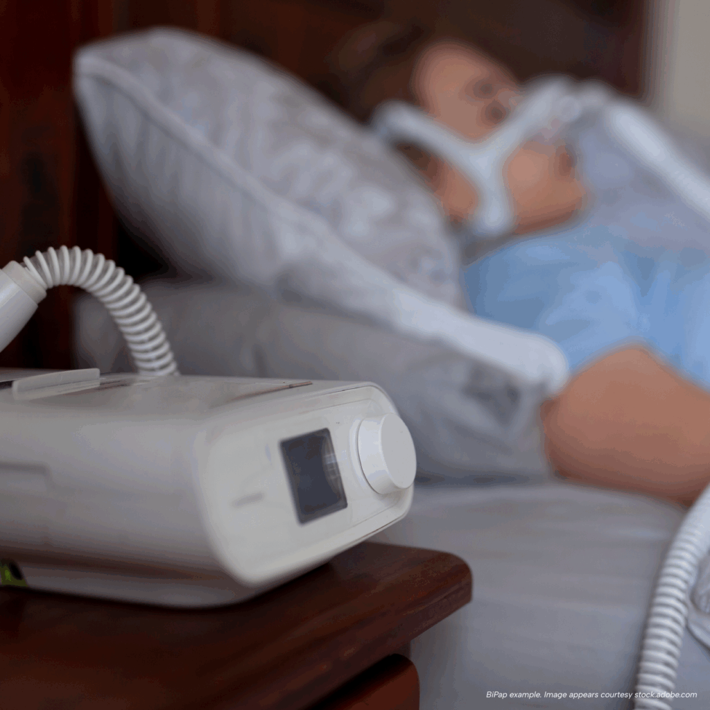 BiPAP machine on a bedside table with tubing connected to a person sleeping while using a nasal mask.