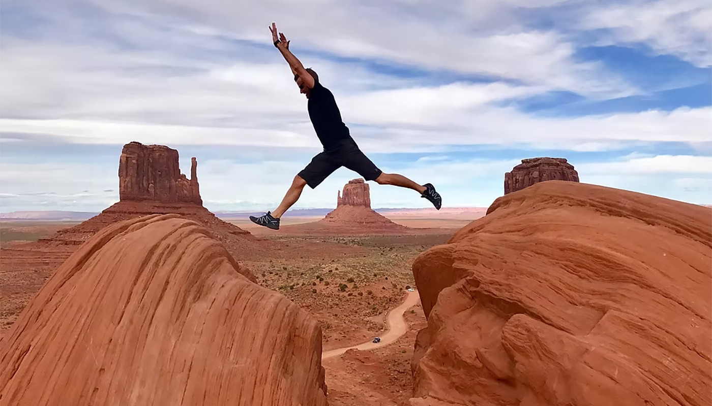 Person jumping between two red rock formations in a desert landscape under a blue sky.
