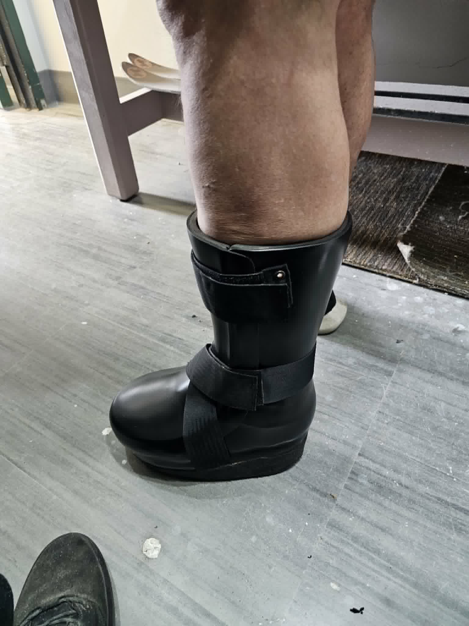 Person standing while wearing a black custom off-loading walker boot that encloses the lower leg and foot, with securing straps and a thick rocker sole.