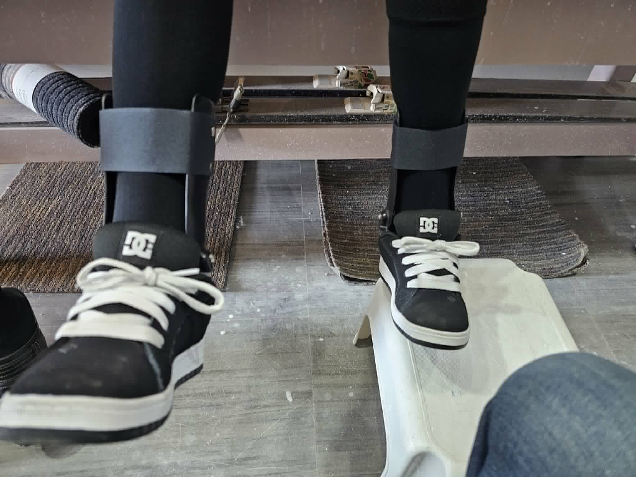 Person seated on an exam table with feet hanging down, wearing bilateral ankle-foot orthoses under socks and black and white sneakers.